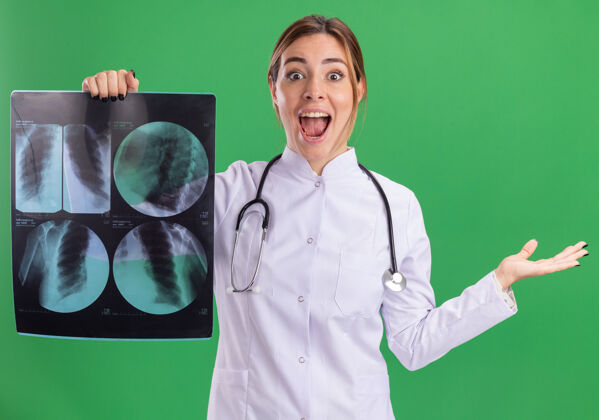 长袍兴奋的年轻女医生穿着医用长袍 手持听诊器 把x射线扩散的手隔离在绿色的墙上兴奋女性抱着