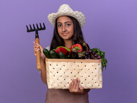 充分穿着围裙 戴着夏帽 拿着装满蔬菜和迷你耙子的小园丁女孩站在紫色的墙上 脸上带着微笑迷你帽子花园