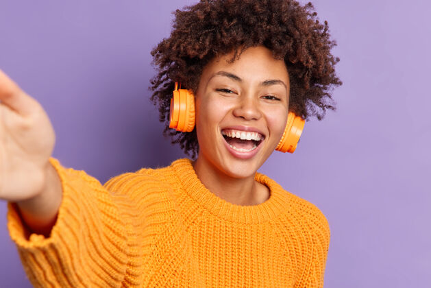 音乐逗乐美丽的非裔美国妇女伸展手臂自拍欢笑愉快地通过无线耳机听音乐穿着橙色针织毛衣的姿势自拍非洲伸展