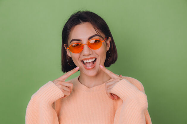 完美年轻时尚的女人穿着休闲的桃色毛衣 戴着橙色眼镜 隔着绿橄榄色的墙壁 积极地微笑着 用食指指着白色的牙齿 复制空间酷女性护理