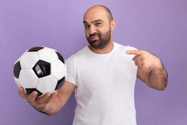 球身穿白色t恤的大胡子男人拿着足球 自信的表情用食指指着站在紫色墙上的一边男人侧线站