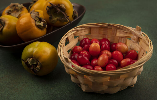 美味顶视图橙色柿子水果在一个碗上与一个桶在绿色的表面上的山茱萸樱桃配料食物甜点