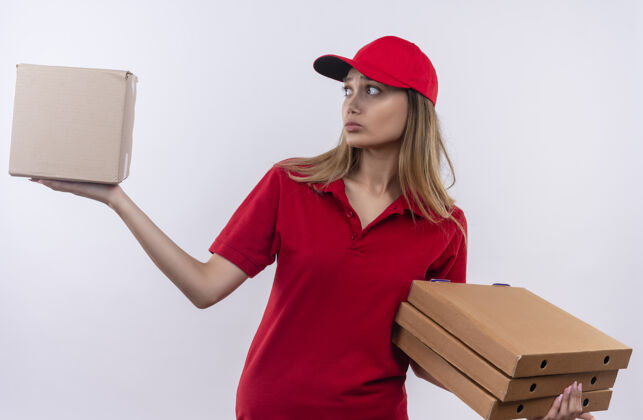 送货困惑的年轻送货员穿着红色制服 戴着帽子 手里拿着披萨盒 看着盒子披萨盒子手