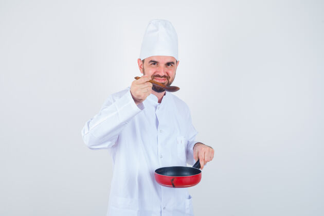 成人年轻的男厨师穿着白色制服 拿着木勺子品尝美食 看上去很愉快正面图微笑美食家服务