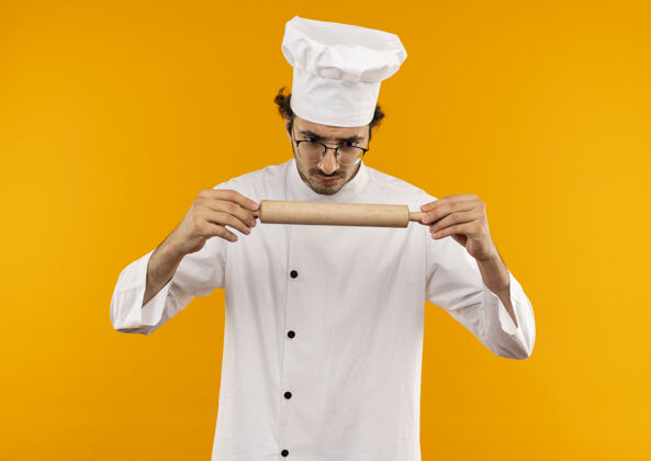 男性困惑的年轻男厨师穿着厨师制服 戴着眼镜拿着擀面杖看着卷厨师别针