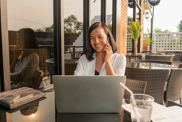 说话一个女商人在咖啡馆里用笔记本电脑和手机交谈的画像女性电脑女商人