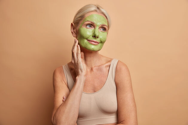 面霜《梦幻中年妇女画像》将绿色面具贴在脸上 若有所思地站在那里 看着远方 在米色的墙上随意地穿上隔离的衣服 接受美容程序成熟美丽润肤霜