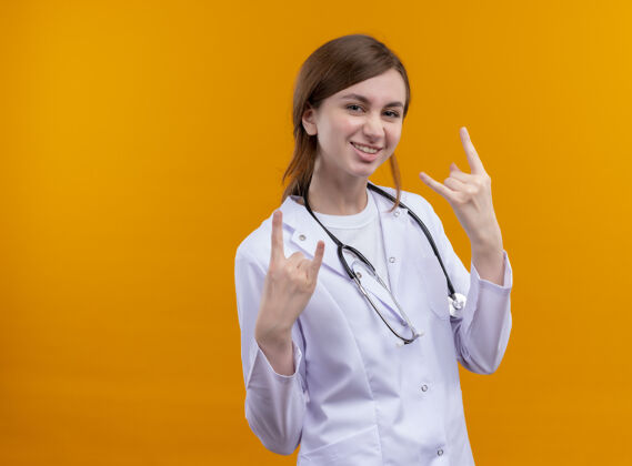 医生微笑的年轻女医生穿着医用长袍和听诊器在隔离的橙色墙壁上做着摇滚标志 还有复制空间听诊器女医学