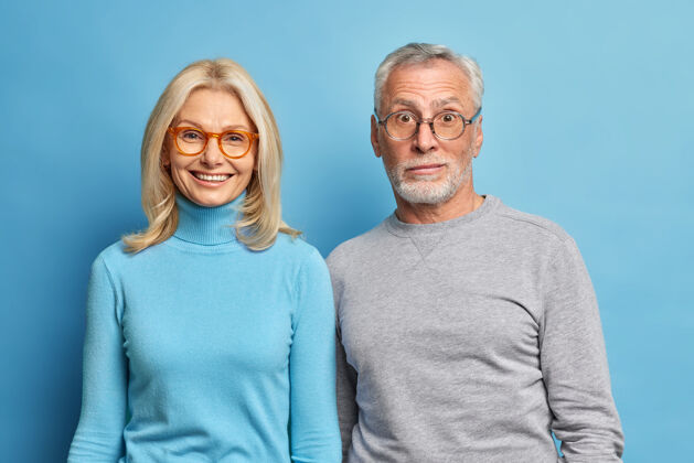 男性惊讶的大胡子男人和中年金发快乐女人的肖像站得很近 穿着休闲套头衫 隔着蓝色的工作室墙眼镜人脸