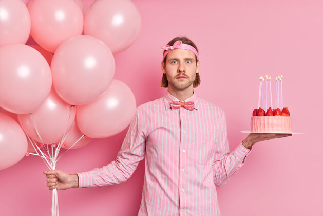 心烦惊喜的男人庆祝生日拿着一堆气球和草莓蛋糕身着正式衬衫领结震惊地看到许多客人在聚会上被隔离在粉红色的墙上正式举行人