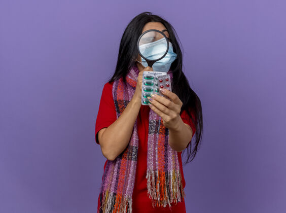 穿戴着口罩和围巾的年轻白种人病女孩伸出一包包胶囊 透过放大镜看着他们 隔离在紫色的墙上 留着复印空间胶囊玻璃包装