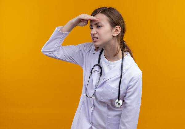 近年轻的女医生穿着医用长袍 手持听诊器 把手放在额头附近 看着隔离的橙色墙上的远处医生手不舒服