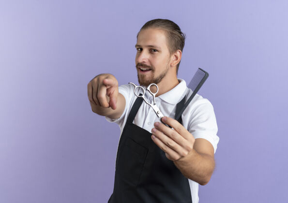理发印象深刻的年轻英俊的理发师穿着制服 伸出梳子和剪刀 指着孤立的紫色与复制空间复制剪刀梳子