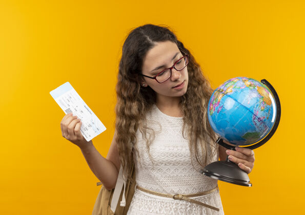 地球仪自信的年轻漂亮的女学生戴着眼镜背着包拿着机票和地球仪看着地球仪孤立在黄色飞机自信年轻