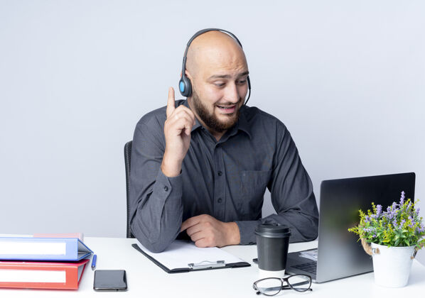 工作令人印象深刻的年轻秃头呼叫中心男子戴着耳机坐在办公桌旁的工作工具看着笔记本电脑和提高手指隔离在白色穿工具电话
