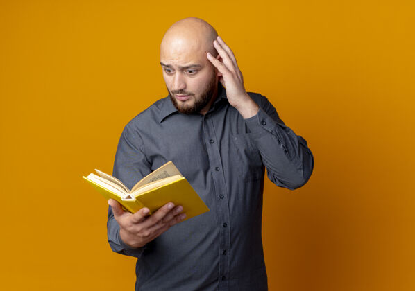 头令人印象深刻的年轻秃头呼叫中心的男子拿着和看的书 手放在头上孤立的橙色与复印空间橙色空间秃头