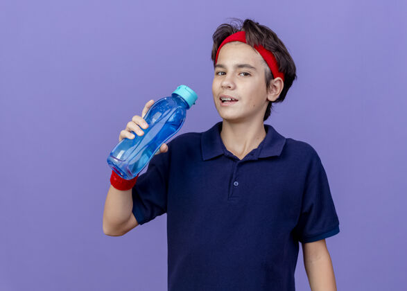 腕带年轻帅气的运动男孩戴着头带和戴着牙套的腕带拿着水瓶看着相机准备喝水在紫色背景上隔离复制空间准备瓶子男孩