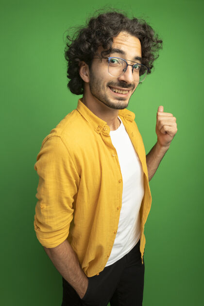 年轻微笑着的年轻英俊的白人男子戴着眼镜站在侧视图中看着相机手放在口袋里指着身后的绿色背景手保持站立