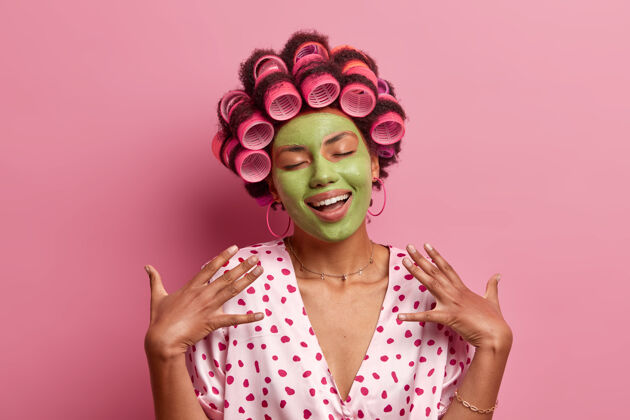 享受快乐的非洲裔美国年轻女子的画像闭上眼睛站着 微笑着 想象着美好的事物 涂上绿色的面膜 卷发器 站在室内人过程长袍