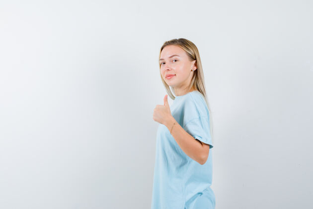 肩膀身着蓝色t恤的金发女孩竖起大拇指 从肩膀看过去 看起来很自信 正面视图肖像情感女性