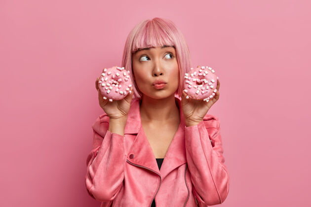 鲍勃梦幻般体贴的亚洲女人拿着两个美味的甜甜圈摆姿势 怀疑是否吃垃圾食品 穿着时髦的粉色夹克 享受甜点人室内粉色假发