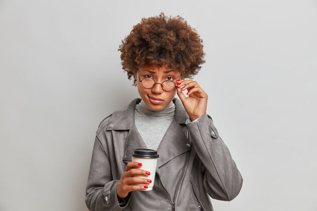 卷发严肃的女学生透过透明眼镜自信地看 专心地听 喝外卖咖啡 穿着灰色夹克眼镜外卖马克杯