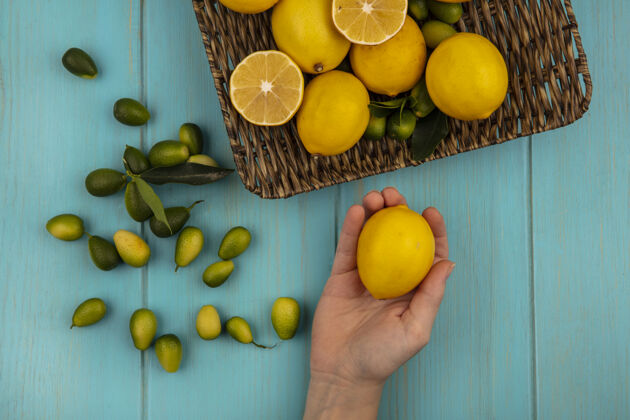 厨房俯视图中的女性手拿着一个柠檬与水果 如金盏花和柠檬柳条托盘上的蓝色木墙柳条切片柑橘