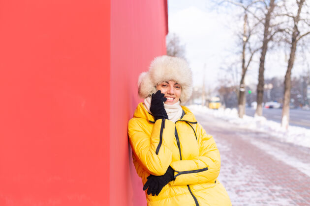 女孩在一个冬天阳光明媚的日子里 一个穿着暖和衣服的红墙背景上的快乐的年轻女子在雪城的人行道上微笑着打电话持有说话通信