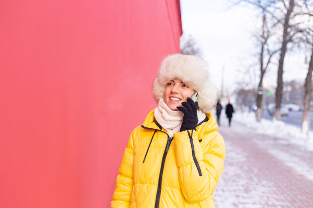 手套在一个冬天阳光明媚的日子里 一个穿着暖和衣服的红墙背景上的快乐的年轻女子在雪城的人行道上微笑着打电话通信微笑帽子