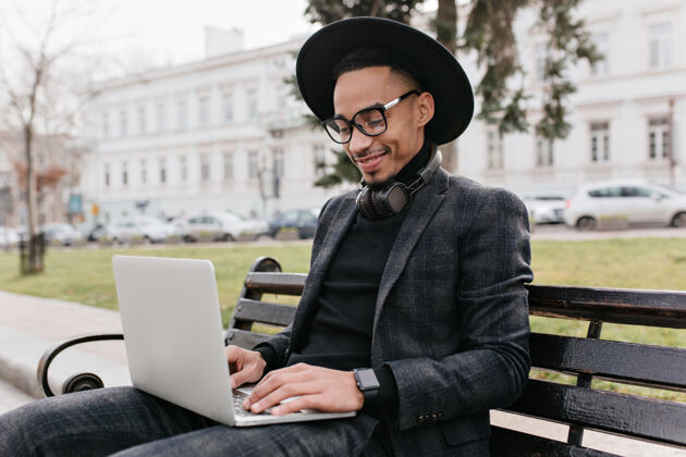 笔记本电脑时髦的国际学生在公园里用笔记本电脑工作微笑的非洲男人在户外用电脑制冷成年人混血儿微笑