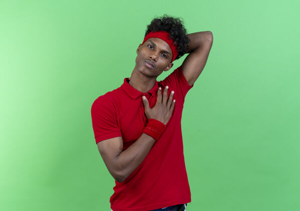 非裔美国人年轻的美国黑人运动男子戴着头带和腕带把一只手放在心上 另一只手放在脖子上隔离在绿色的墙上运动墙头带