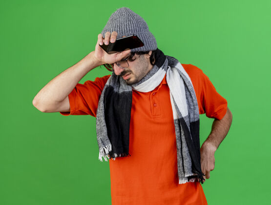 人那个戴着眼镜 戴着冬天的帽子 戴着围巾 拿着手机 拿着餐巾 手放在头上 与世隔绝地站在绿色墙壁上的病痛的年轻人人电话保持