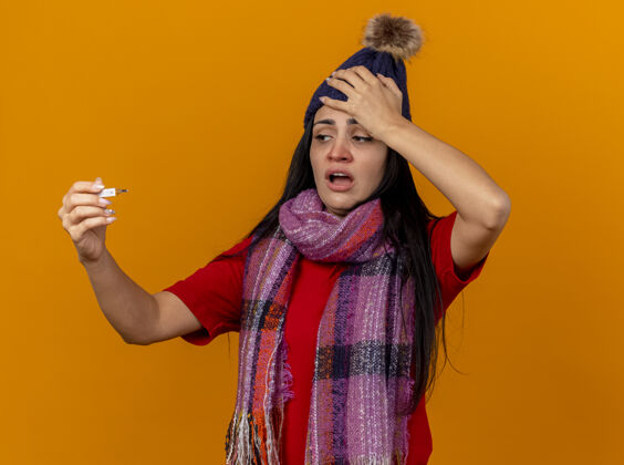 抱着那个戴着冬日帽子 戴着围巾 手放在隔离在橘色墙上的头上 看着温度计的年轻生病的女人冬天体温计围巾