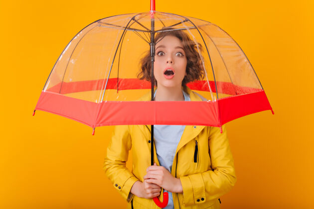 情绪热情的卷发女孩站在阳伞下的特写照片穿着雨衣打伞的心烦意乱的女模特的室内照片肖像姿势女人
