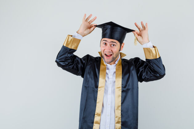 毕业年轻的男性手拿手指在他的帽子上 穿着研究生制服 看起来很高兴前视图休闲帽子肖像
