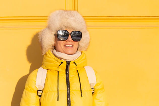 太阳镜在阳光明媚的日子里 一个戴着温暖的俄罗斯西伯利亚帽子的快乐女人的肖像画 她在冬天穿着雪白的zabas 对着黄色的墙壁微笑着手套自拍漂亮
