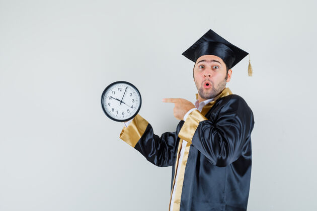 毕业年轻人指着墙上的挂钟 穿着研究生制服 看上去很惊讶男人时钟休闲