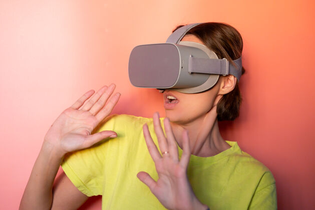 人在粉红色橙色背景的工作室里 戴着虚拟现实眼镜的女人的情感肖像娱乐数码耳机