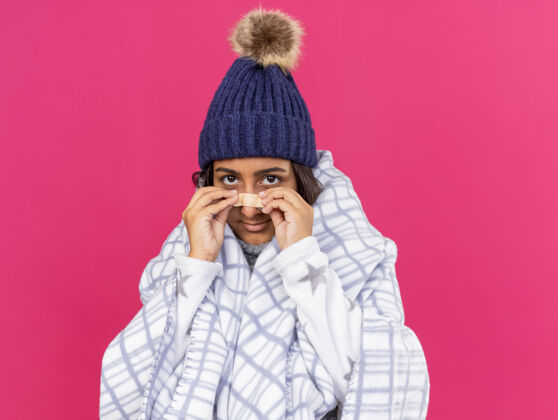 年轻可爱的生病的小女孩戴着冬天的帽子 围巾用格子布包着 鼻子上贴着膏药 背景是粉色的围巾请格子布