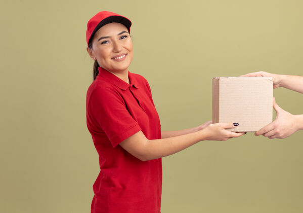 制服身着红色制服 头戴鸭舌帽的年轻女送货员站在绿色墙壁上 微笑着向顾客赠送纸板箱帽子女人送货