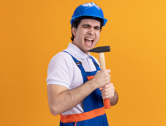 抱着年轻的建筑工人穿着建筑制服 戴着安全帽 拿着锤子作为麦克风 站在橙色的墙上 情绪激动 快乐地歌唱着男人麦克风安全