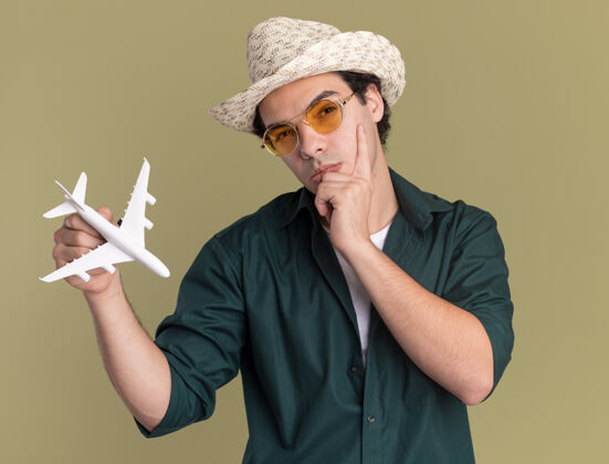 戴着一个穿着绿色衬衫 戴着夏天帽子 戴着眼镜 拿着玩具飞机的年轻人站在绿色的墙上沉思地看着前方飞机年轻人站着