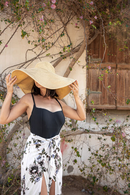 年轻一个年轻的女人躲在一个大草帽下躲避太阳 草帽靠近一堵墙 墙上缠绕着一棵正在开花的树的干树枝热热带女人