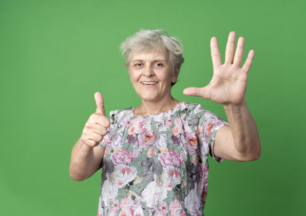 绿微笑的老妇人举手竖起大拇指孤立地站在绿色的墙上举起来老人手