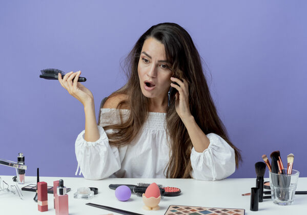 壁板震惊的美女拿着化妆工具坐在桌边拿着梳子讲电话看着紫色墙上孤立的一面抱着坐着工具
