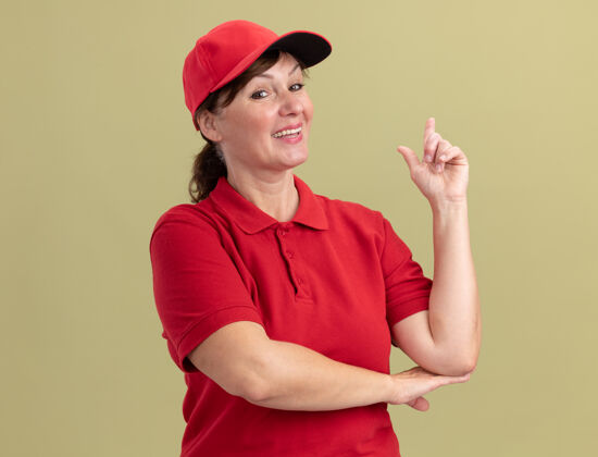 目录身着红色制服 头戴鸭舌帽的中年女送货员站在绿色的墙边 微笑着自信地展示着有新创意的食指分娩中间新的