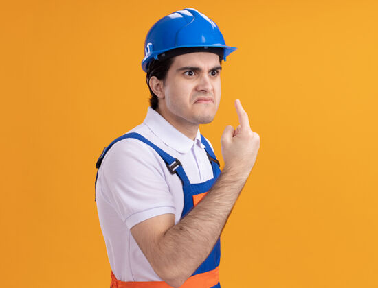头盔年轻的建筑工人穿着建筑制服 戴着安全帽 愤怒的脸上露出食指警告站在橙色的墙上手指建筑脸