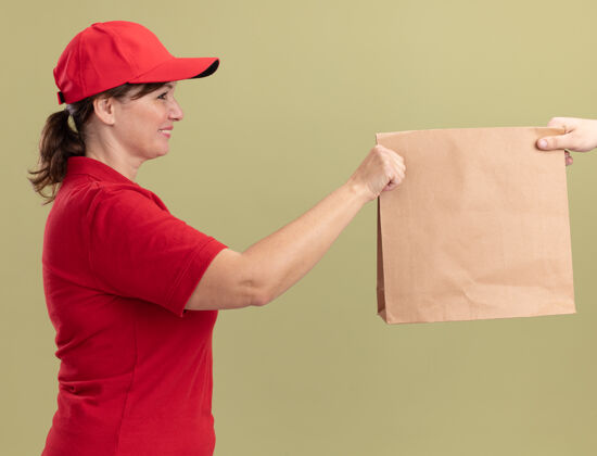 纸身着红色制服 头戴帽子的中年送货员站在绿色的墙上 微笑着向顾客赠送纸质包装包装制服年龄