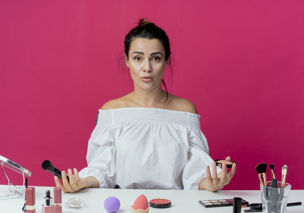 工具惊讶美丽的女孩坐在桌旁 拿着化妆工具 拿着粉和化妆刷隔离在粉红色的墙上桌子惊喜画笔