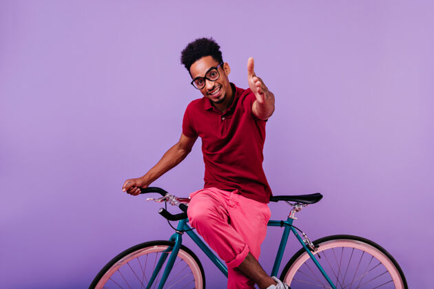 室内感兴趣的非洲男模摆姿势戴眼镜的时髦黑人男孩坐在蓝色自行车上快乐兴奋黑色